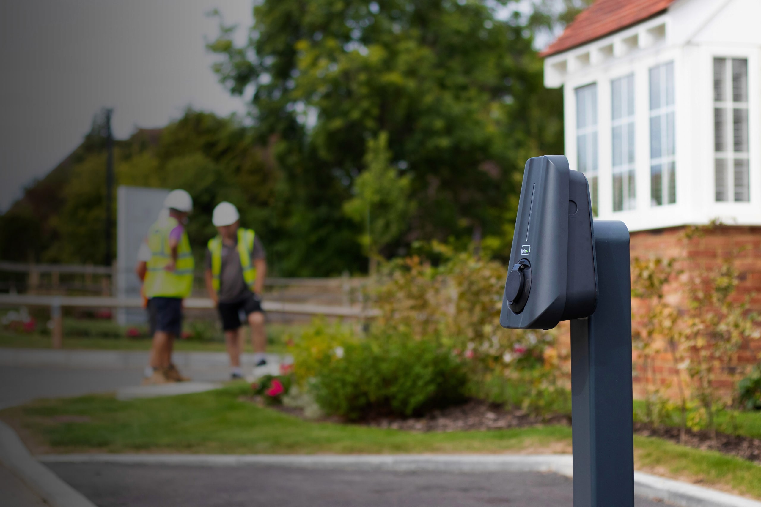 Brim Smart EV Charger hero image, a portrait style shot of the Brim Charger mounted to a post with blurred new build residential site and three trades people in high vis jackets in the background.
